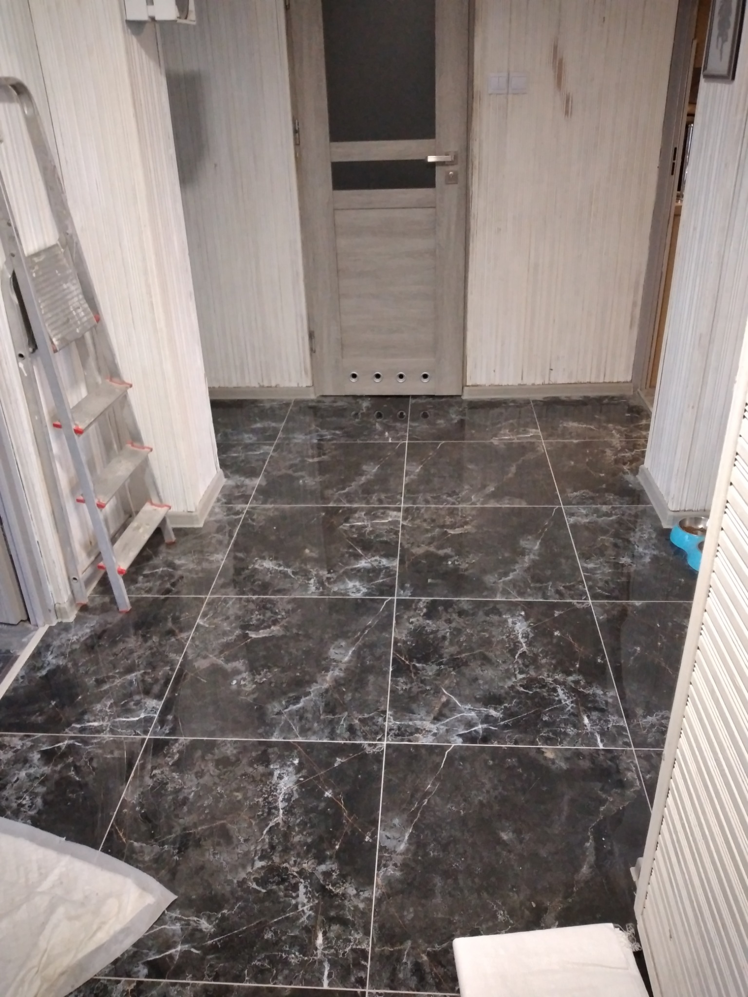 Ciemne, marmurkowe płytki podłogowe w trakcie układania, widoczna drabina malarska i fragment niebieskiej miski w rogu pomieszczenia.