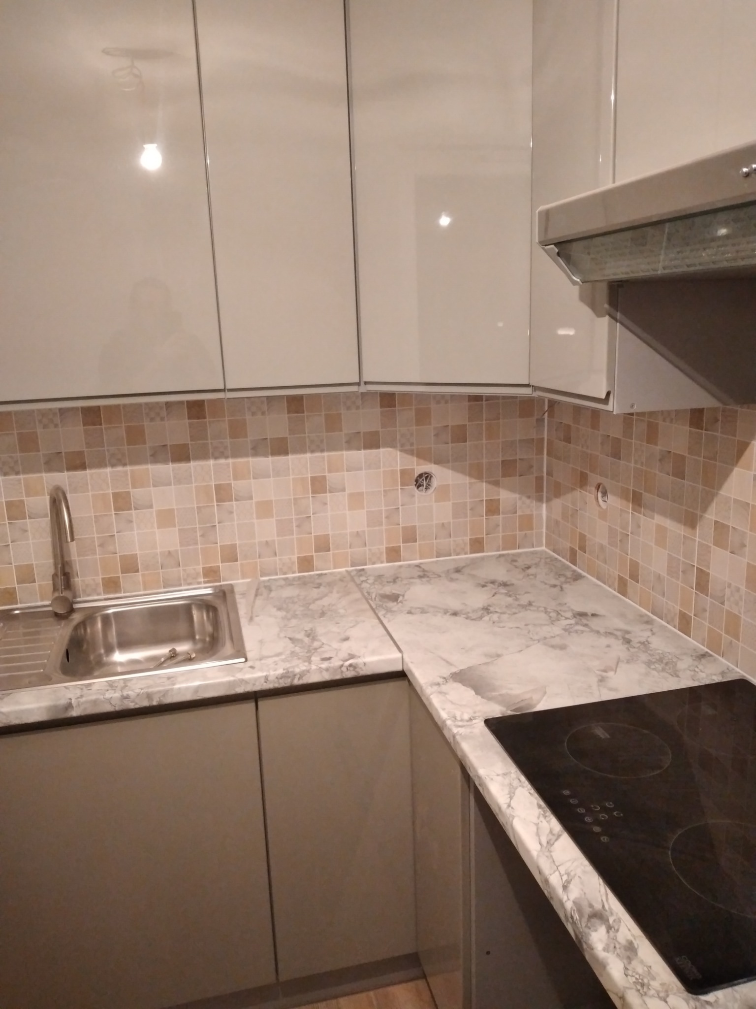 Wykończona kuchnia z błyszczącymi, kremowymi szafkami górnymi, szarymi szafkami dolnymi, mozaiką na ścianie, blatem imitującym marmur i czarną płytą indukcyjną