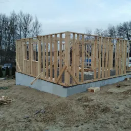 Surowy szkielet drewnianej konstrukcji domu na betonowym fundamencie, widoczne deski i elementy łączeniowe, teren budowy w tle z drzewami i przyczepą kempingową.