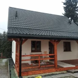 Niewielki dom z drewnianym tarasem i ciemną, metalową dachówką, sfotografowany w pochmurny dzień podczas opadów deszczu lub śniegu; widoczne drewniane słupy podtrzymujące dach tarasu i elementy...