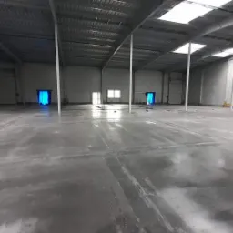 Wnętrze nowo wybudowanej hali z betonową posadzką, widoczne białe słupy konstrukcyjne, świetliki w dachu i dwa wejścia zabezpieczone niebieską folią.