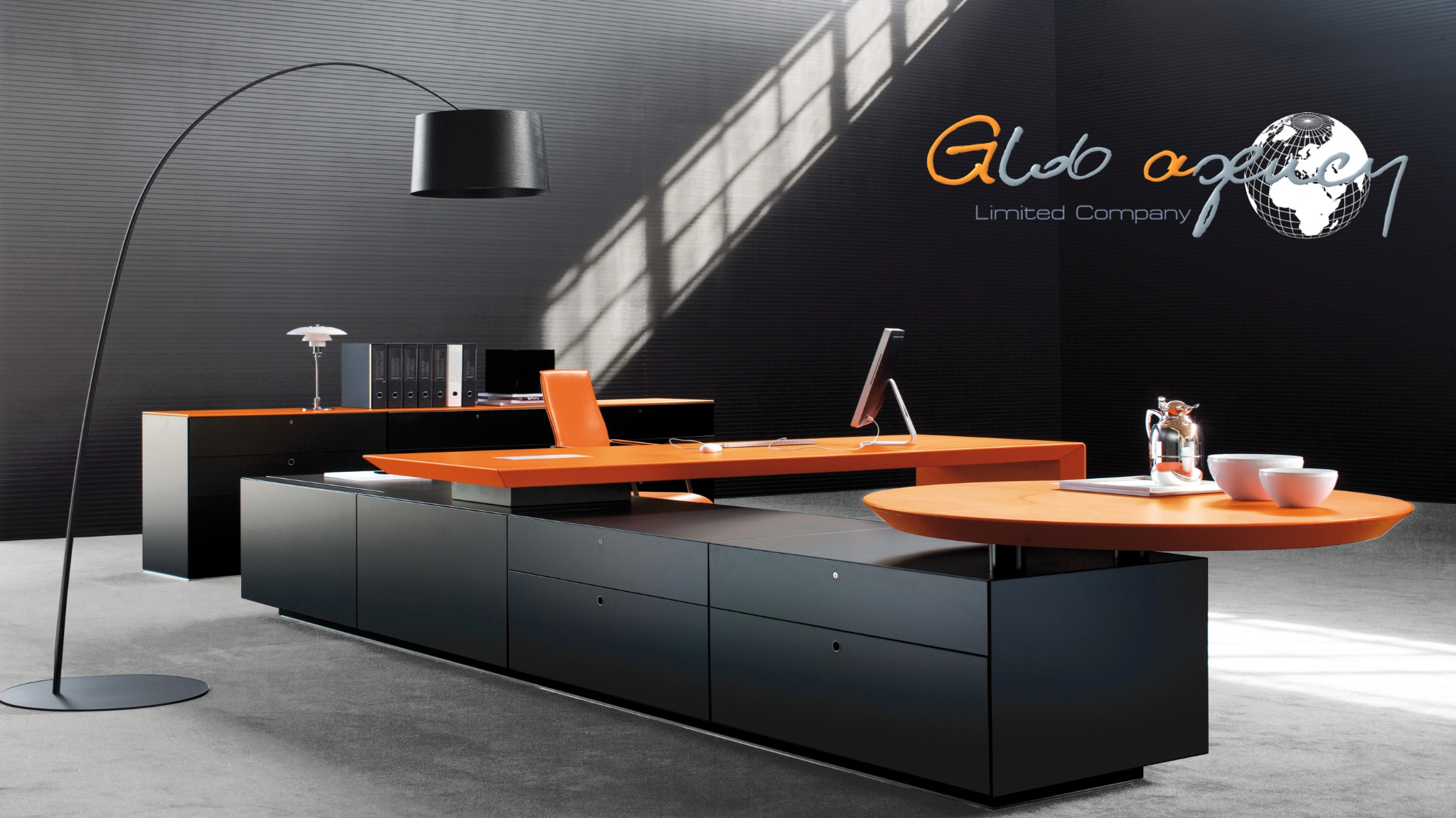 Nowoczesne biuro z czarnymi meblami i pomarańczowymi blatami, oświetlone lampą łukową i światłem słonecznym wpadającym przez okno, na ścianie logo firmy 'Glob agency'.