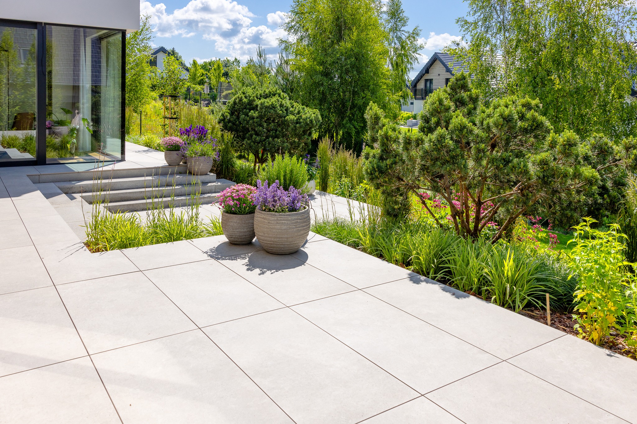 Nowoczesny taras z jasnych, wielkoformatowych płyt, donice z kwiatami, schody i bujna zieleń w tle. Minimalistyczny design i harmonia z naturą.