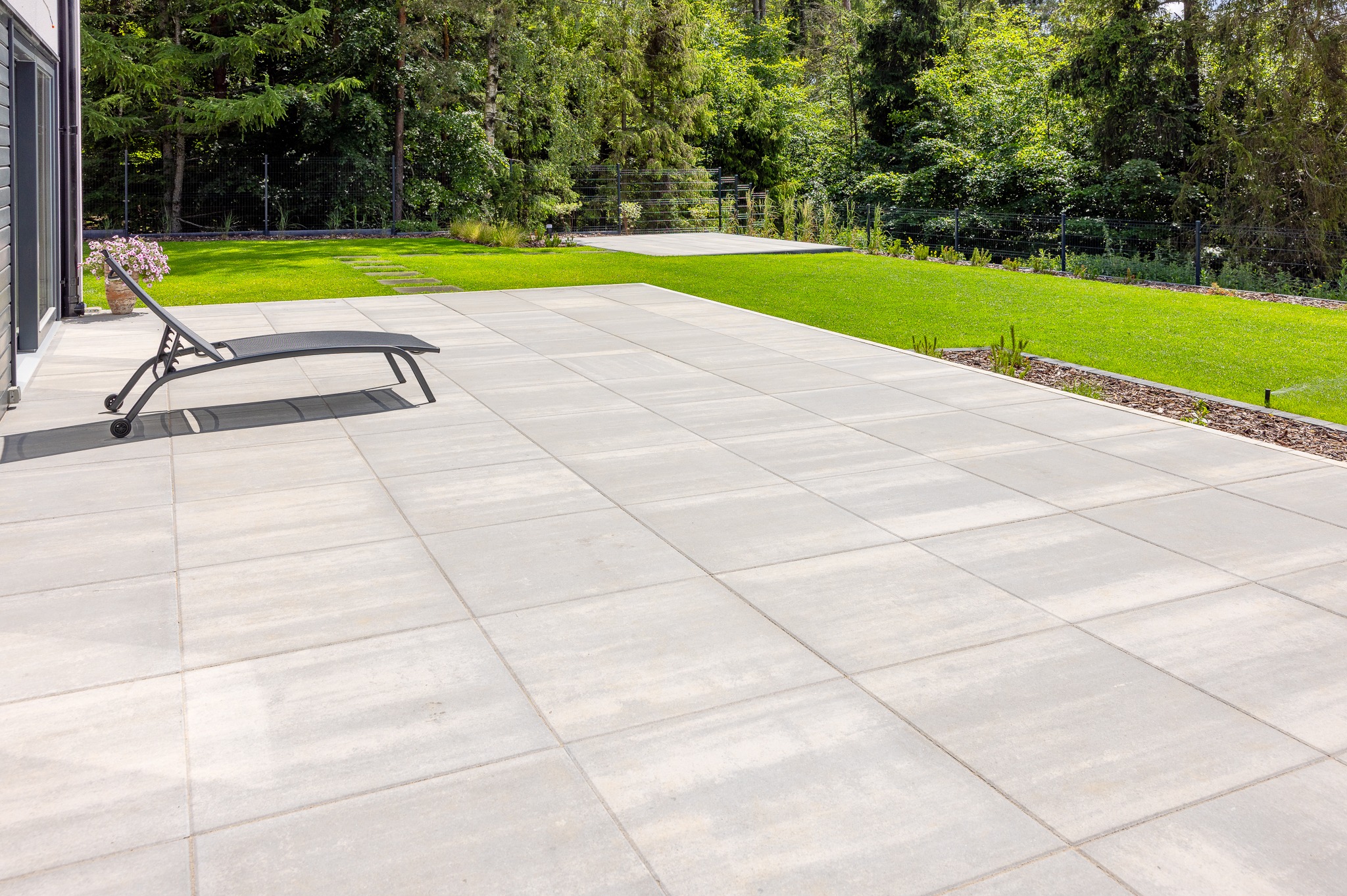 Jasnoszara, duża powierzchnia z kostki brukowej na zewnątrz, z leżakiem i zielonym trawnikiem w tle. Widoczne drzewa i krzewy. Minimalistyczny design.