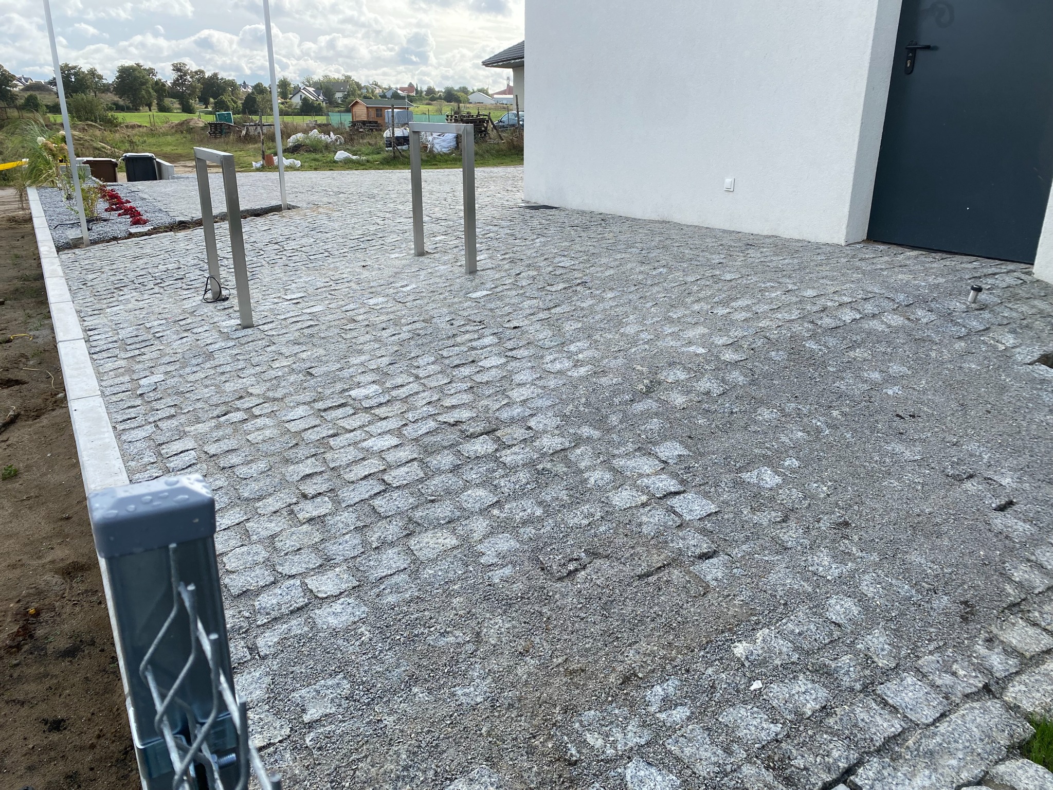 Szara kostka granitowa ułożona na podjeździe, widoczne słupki i fragment ogrodzenia. Tło stanowi budynek z szarą stolarką i zieleń. Całość w dziennym oświetleniu.