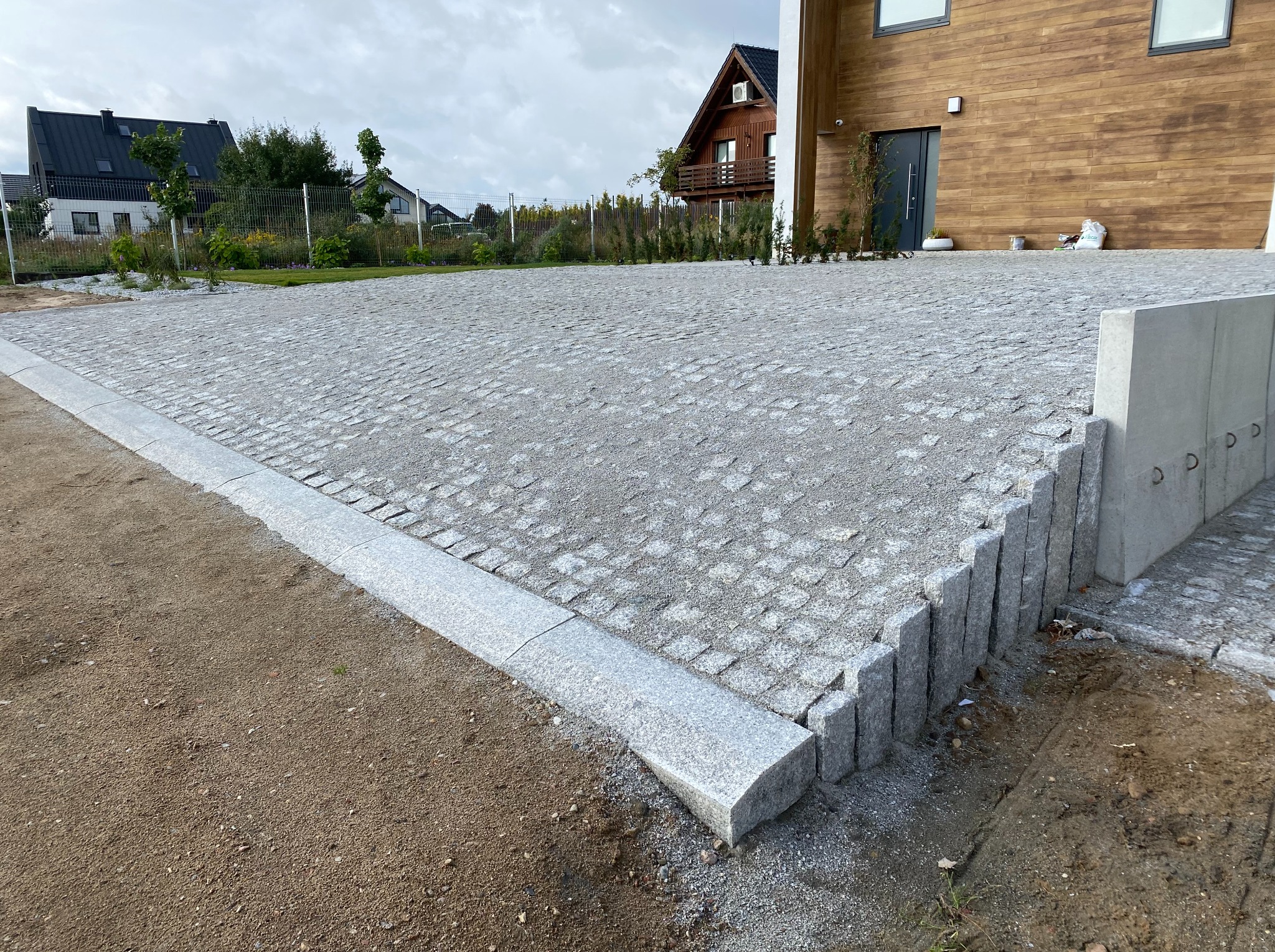 Ułożona kostka granitowa na podjeździe domu jednorodzinnego, z widocznymi obrzeżami i elementami wykończeniowymi. Tło stanowi budynek z drewnianą elewacją.