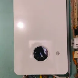 Biały piec gazowy Junkers zamontowany na pomalowanej na zielono ścianie, widoczne rury miedziane i żółty przewód gazowy, oraz szary przewód odprowadzający spaliny.