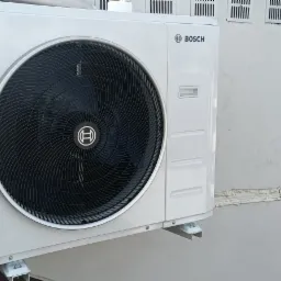 Zamontowana jednostka zewnętrzna klimatyzacji Bosch na szarej ścianie budynku. Widoczne elementy montażowe i fragment instalacji. W tle zabudowa miejska.