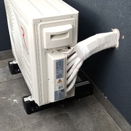 SERWIS GAZOWYCH URZĄDZEŃ GRZEWCZYCH -PAWEŁ ROKOSZ - Biały klimatyzator Rotenso zamontowany na zewnątrz budynku, z widocznymi rurami owiniętymi taśmą, prowadzącymi przez otwór w szarej ścianie. Widok z dołu.