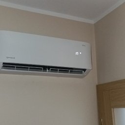 SERWIS GAZOWYCH URZĄDZEŃ GRZEWCZYCH -PAWEŁ ROKOSZ - Biały klimatyzator Rotenso zamontowany na beżowej ścianie w pomieszczeniu. Widoczny fragment okna z żaluzjami i drzwi z drewnianą ościeżnicą. Na klimatyzatorze wyświetlana temperatura 17 stopni.
