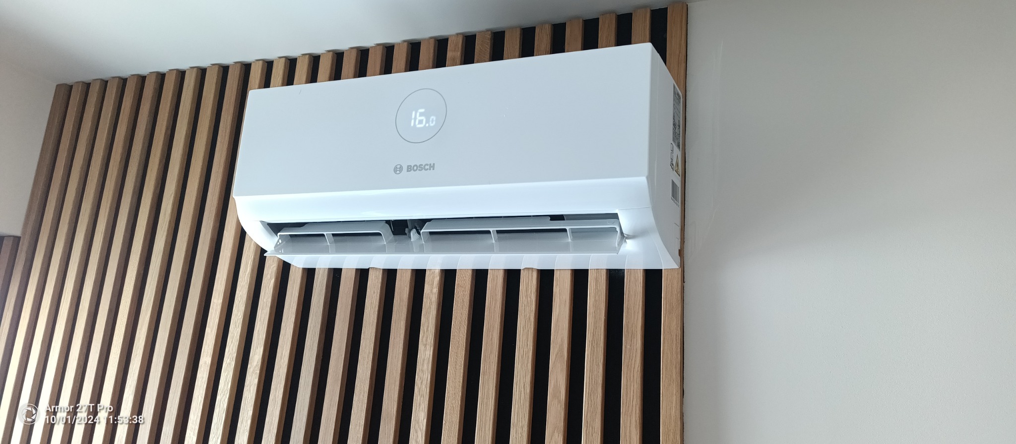 Biały klimatyzator Bosch zamontowany na ścianie z drewnianymi lamelami. Widoczny wyświetlacz z temperaturą 16 stopni. Świeżo po montażu, zabezpieczony taśmą.