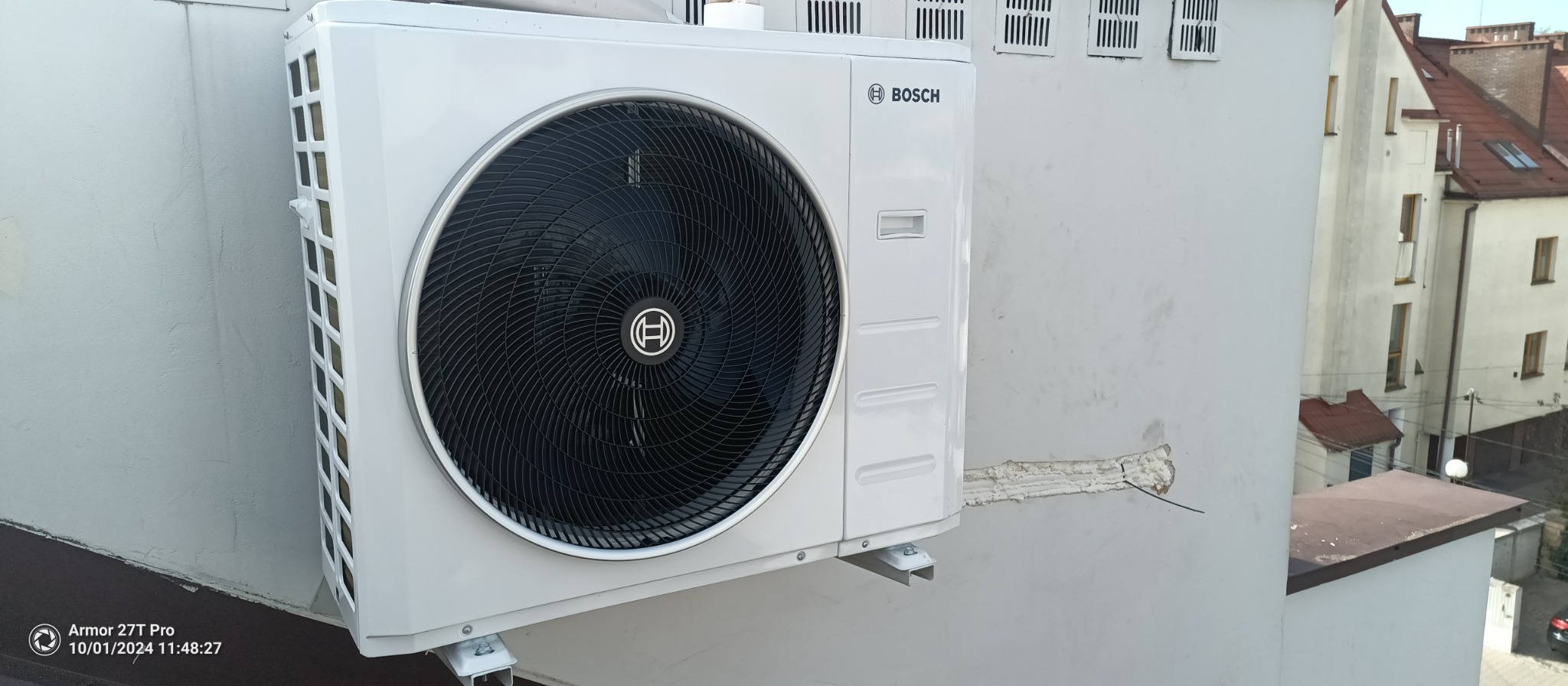Zamontowana jednostka zewnętrzna klimatyzacji Bosch na szarej ścianie budynku. Widoczne elementy montażowe i fragment instalacji. W tle zabudowa miejska.