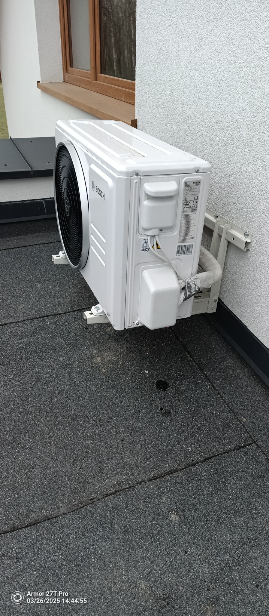 Zamontowana jednostka zewnętrzna klimatyzatora Bosch na elewacji budynku. Widoczne elementy montażowe i instalacja. Szara powierzchnia dachu płaskiego w tle.