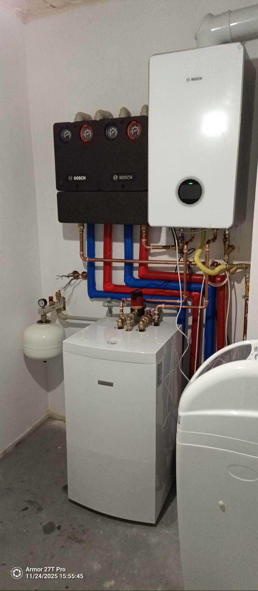 Nowoczesny, biały piec gazowy Bosch z czarnym modułem sterującym i instalacją z rur miedzianych i izolowanych na niebiesko-czerwono, w kotłowni. Widoczny zbiornik wyrównawczy.