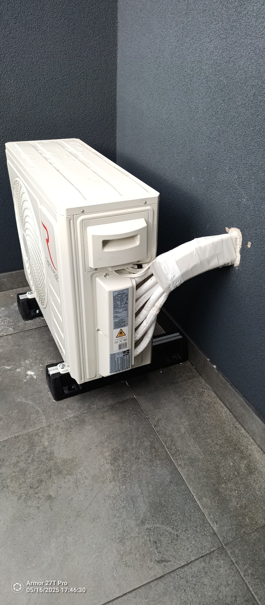 Biały klimatyzator Rotenso zamontowany na zewnątrz budynku, z widocznymi rurami owiniętymi taśmą, prowadzącymi przez otwór w szarej ścianie. Widok z dołu.