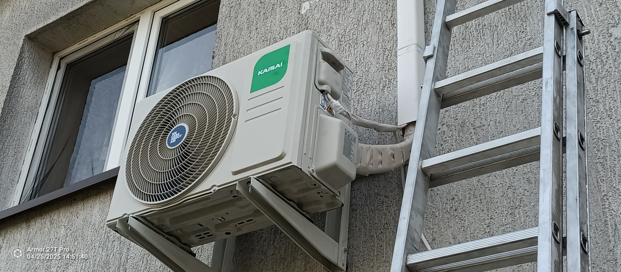 Zamontowany klimatyzator Kaisai na szarej elewacji budynku, widoczna drabina oparta o ścianę. Instalacja zabezpieczona izolacją. Ujęcie z dołu.