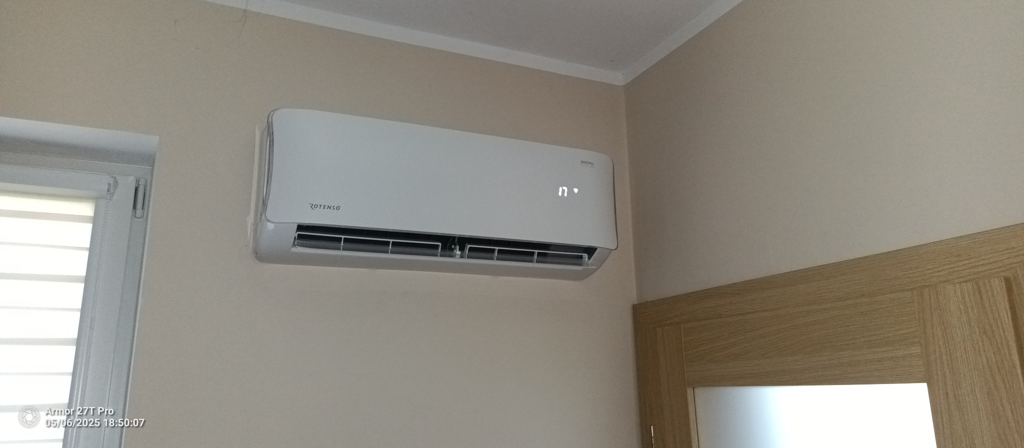 Biały klimatyzator Rotenso zamontowany na beżowej ścianie w pomieszczeniu. Widoczny fragment okna z żaluzjami i drzwi z drewnianą ościeżnicą. Na klimatyzatorze wyświetlana temperatura 17 stopni.