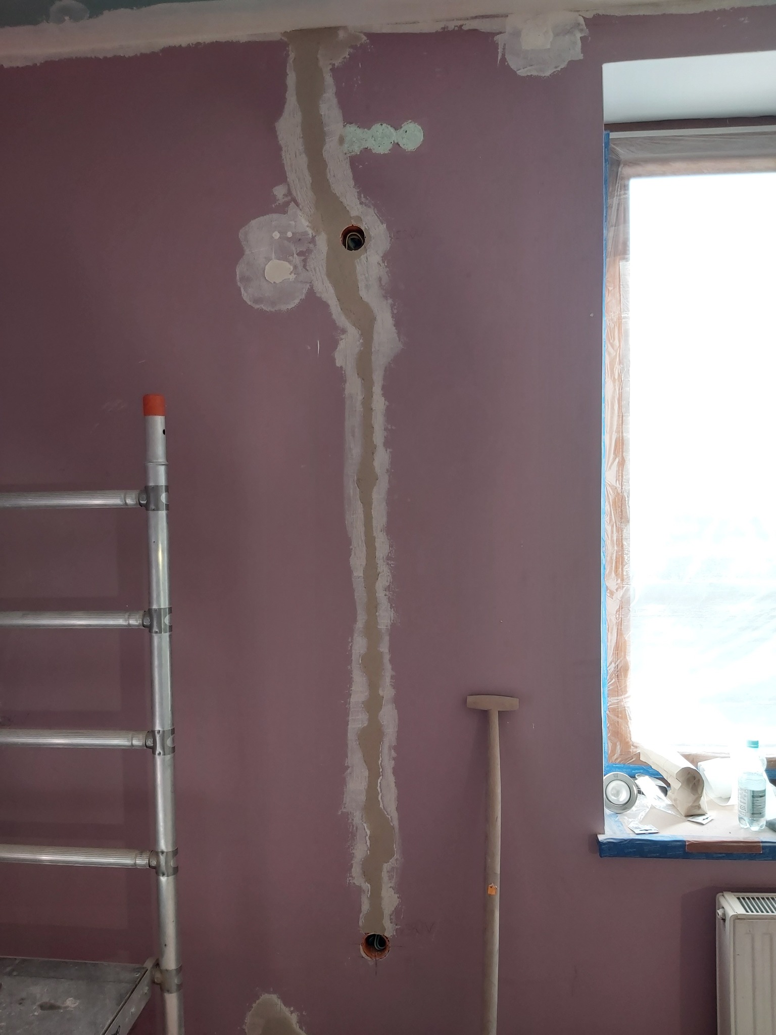 Fioletowa ściana w trakcie remontu z wyżłobionym pionowym kanałem na przewody elektryczne i puszkami instalacyjnymi, częściowo wypełnionym gipsem, drabina aluminiowa z lewej strony, okno...