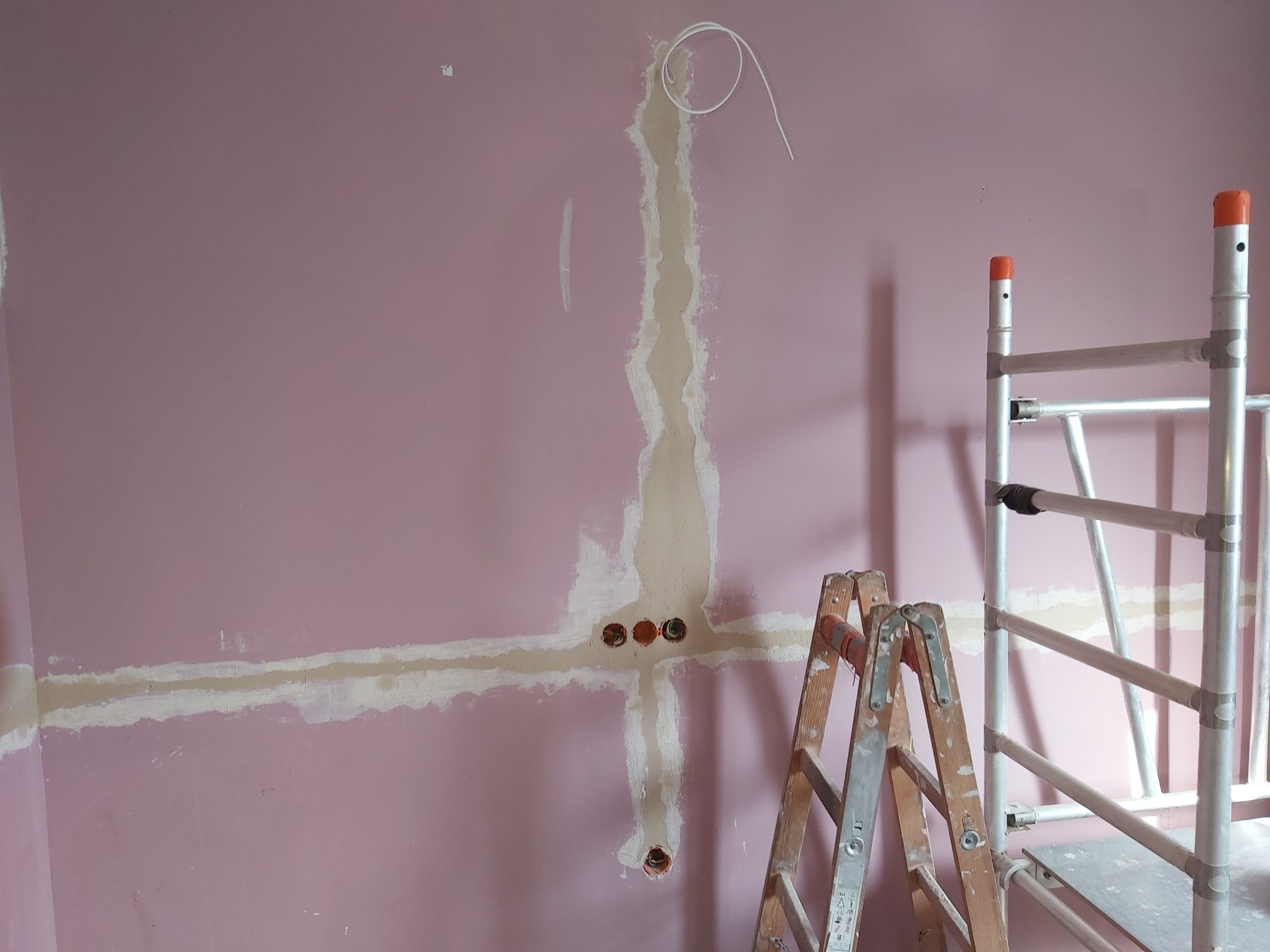 Różowa ściana w trakcie przygotowania do prac elektrycznych: widoczne bruzdy wypełnione gipsem, otwory pod gniazdka, wystający przewód, drabina drewniana i aluminiowy ruszt.