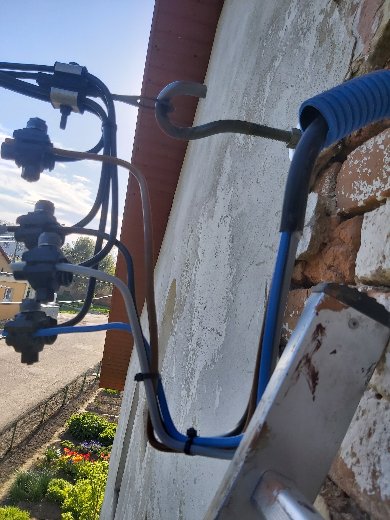 Kable elektryczne przymocowane do ściany budynku z widoczną drabiną, niebieską rurką karbowaną i elementami łączeniowymi, z fragmentem ogrodu i ulicy w tle.