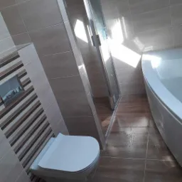 Nowoczesna łazienka po remoncie: beżowe płytki ścienne z dekoracyjnym pasem, brązowe płytki podłogowe imitujące drewno, biała wisząca toaleta i kabina prysznicowa z transparentnym szkłem.