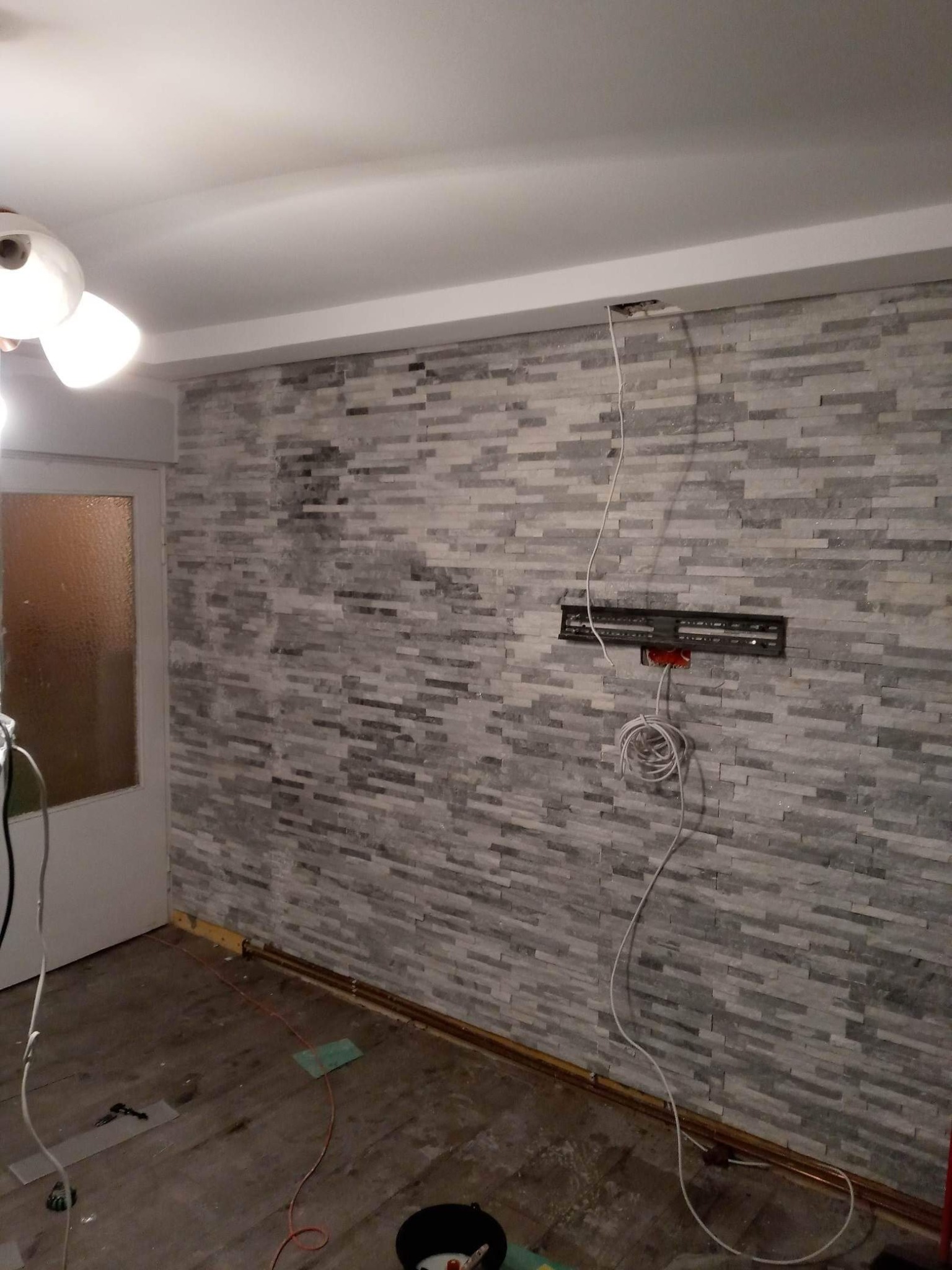 Ściana wykończona szarą okładziną kamienną w trakcie montażu, widoczne kable, puszka elektryczna i listwa przypodłogowa, podłoga częściowo zabezpieczona folią.
