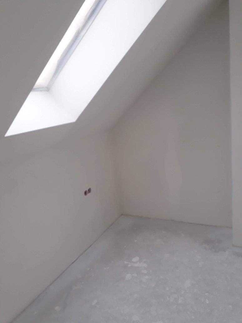 Poddasze z oknem dachowym przed wykończeniem: surowe ściany, widoczne gniazdka elektryczne, szary betonowy podkład podłogowy.