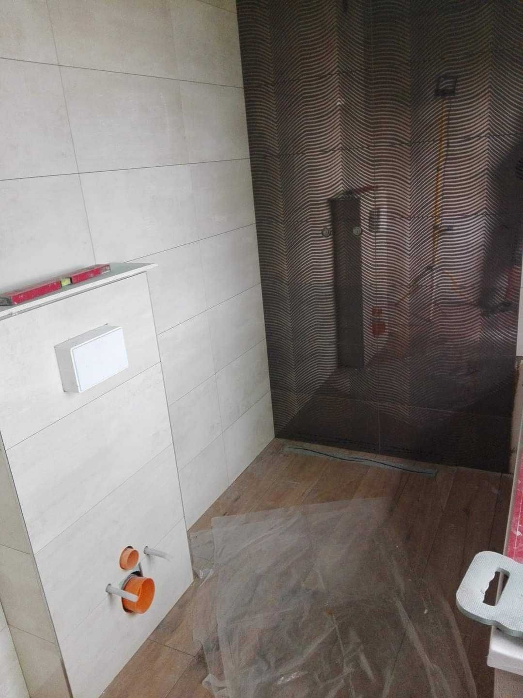 Łazienka w trakcie wykańczania: widoczne płytki na ścianach, podwieszana toaleta z instalacją, oraz fragment kabiny prysznicowej z dekoracyjnymi płytkami. Podłoga częściowo zabezpieczona folią.