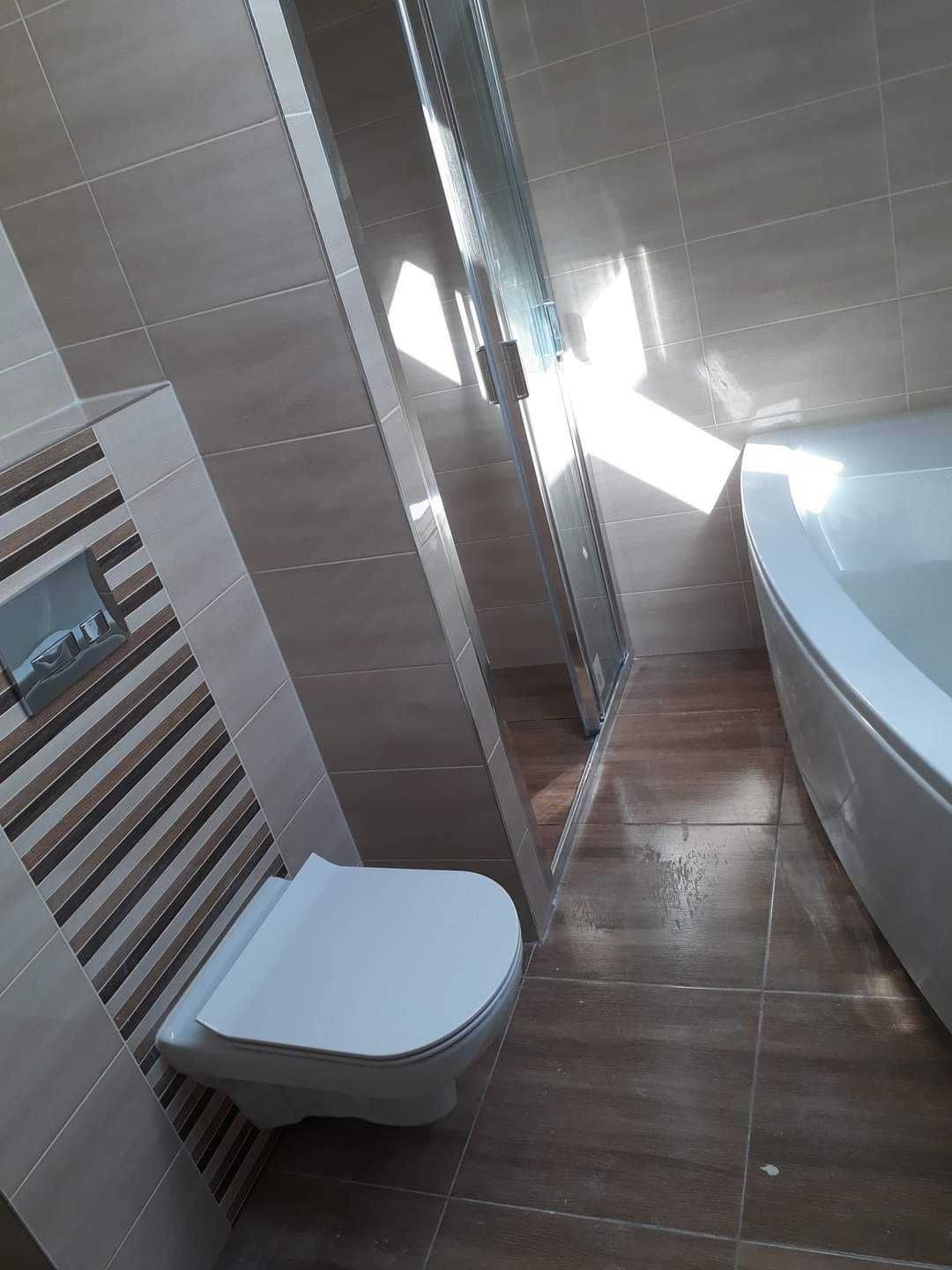 Nowoczesna łazienka po remoncie: beżowe płytki ścienne z dekoracyjnym pasem, brązowe płytki podłogowe imitujące drewno, biała wisząca toaleta i kabina prysznicowa z transparentnym szkłem.