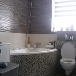 Nowoczesna łazienka z wanną obudowaną mozaiką, płytkami imitującymi fale na ścianach, szarą podłogą i białą armaturą. Widoczny fragment pralki i kosz na pranie.