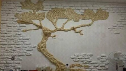 Dekoracyjna ściana z motywem drzewa wykonana z wypukłych elementów imitujących cegły i korę drzewa, częściowo pomalowana na biało, z widocznymi niedoskonałościami w wykończeniu.
