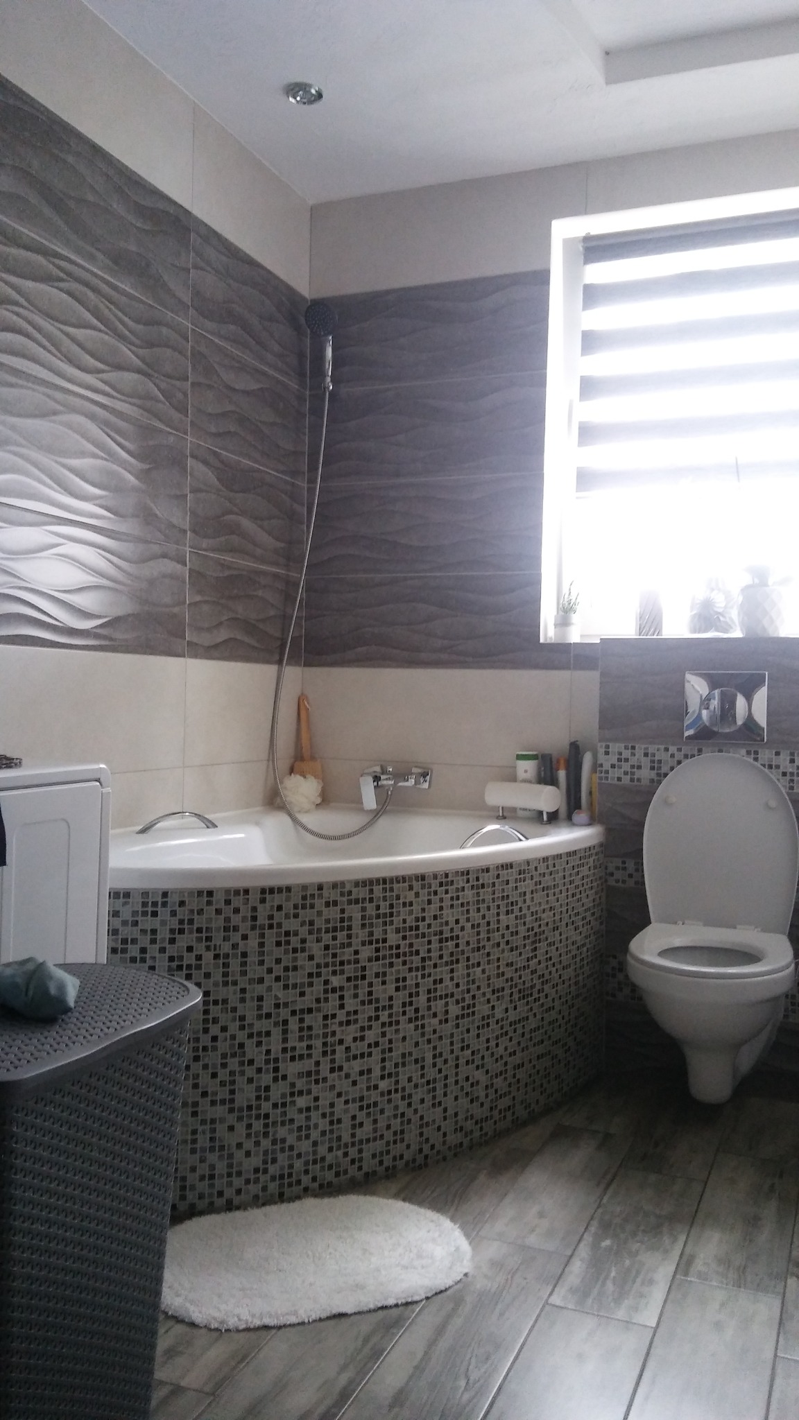 Nowoczesna łazienka z wanną obudowaną mozaiką, płytkami imitującymi fale na ścianach, szarą podłogą i białą armaturą. Widoczny fragment pralki i kosz na pranie.