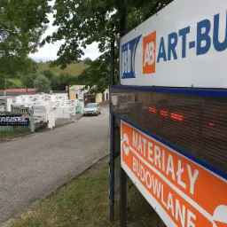 Widok z zewnątrz na skład z materiałami budowlanymi, z tablicą informacyjną i stosem płyt ociepleniowych w tle, widoczny samochód osobowy.