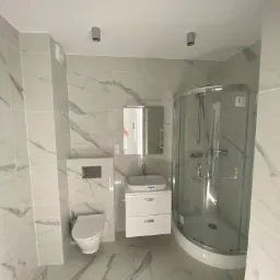 Nowoczesna łazienka wykończona w białych płytkach imitujących marmur, z kabiną prysznicową z zaokrąglonym frontem, podwieszaną toaletą i szafką z umywalką.