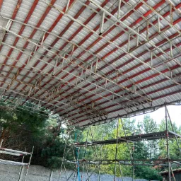 Widok od dołu na konstrukcję dachu z blachy trapezowej, wspartą na metalowych dźwigarach w kolorze czerwonym, z widocznymi rusztowaniami i zielenią drzew w tle.