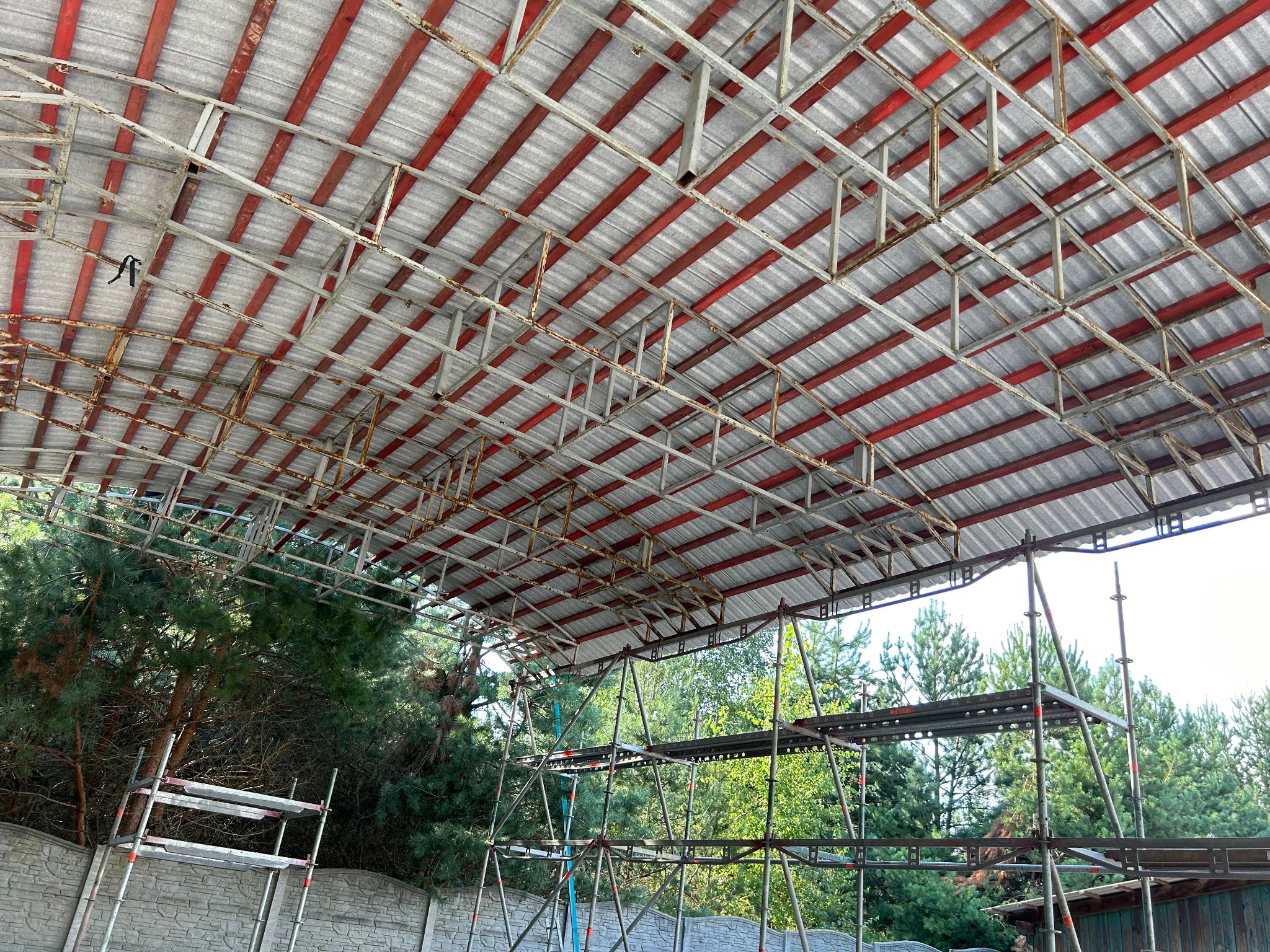 Widok od dołu na konstrukcję dachu z blachy trapezowej, wspartą na metalowych dźwigarach w kolorze czerwonym, z widocznymi rusztowaniami i zielenią drzew w tle.
