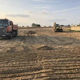 Pomarańczowa ciężarówka Scania i żółta koparka na przygotowanym terenie budowlanym z widocznymi śladami opon i świeżo usypaną ziemią.