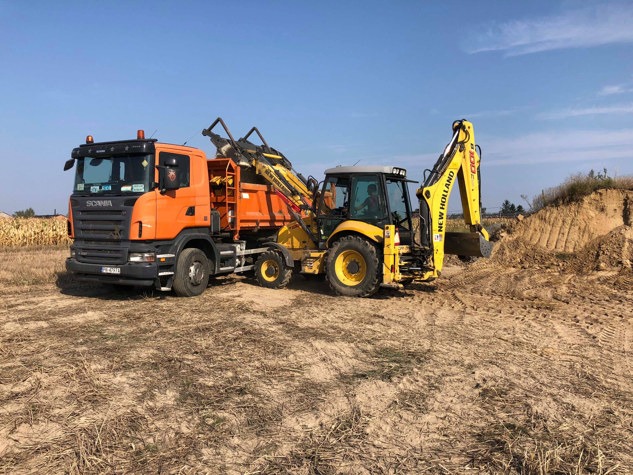 Pomarańczowa ciężarówka Scania z załadowaną ziemią obok żółtej koparko-ładowarki New Holland podczas prac ziemnych na placu budowy w słoneczny dzień.