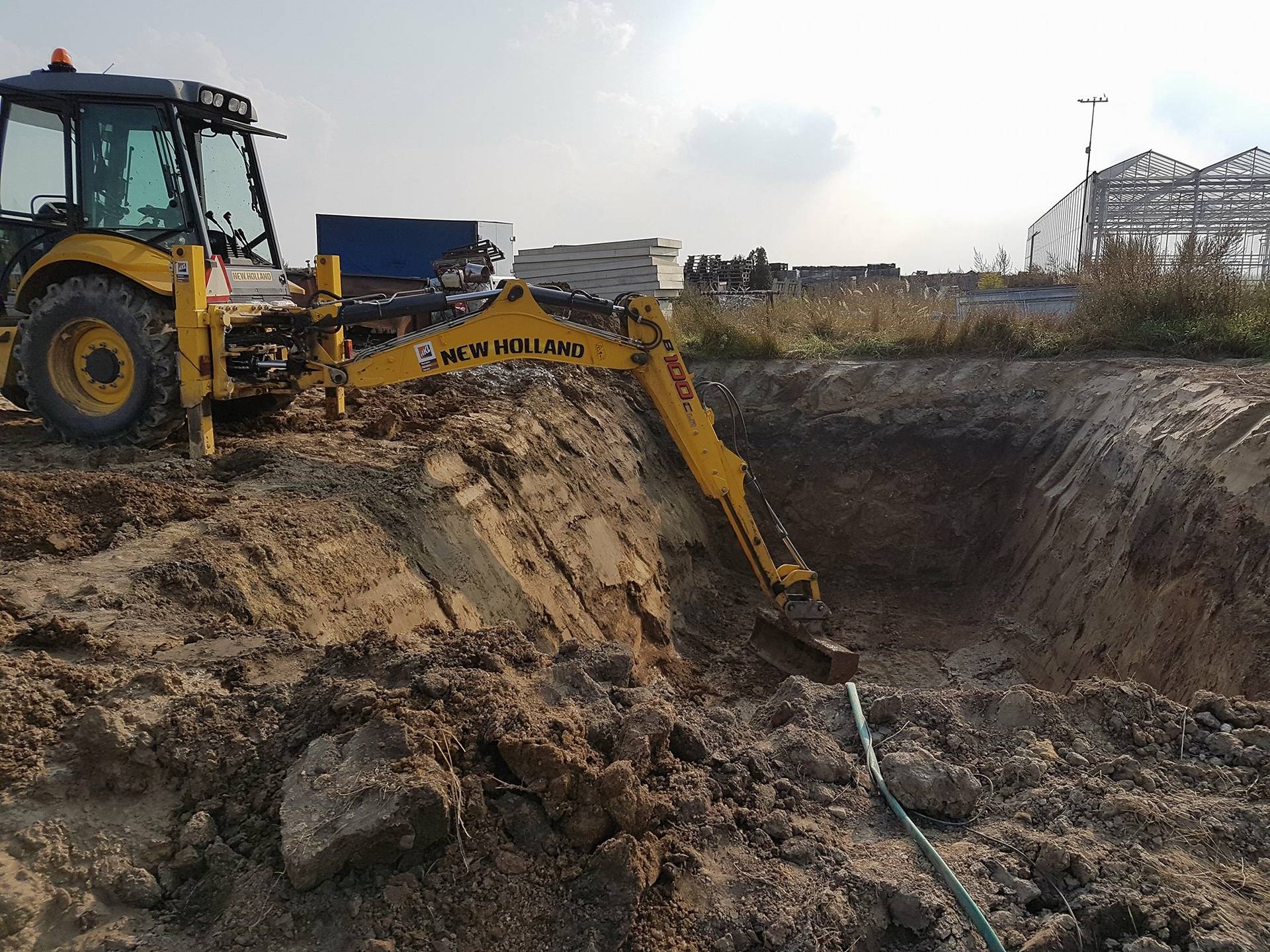 Żółta koparko-ładowarka New Holland wykonuje wykop w ziemi, widoczny fragment zielonego węża, w tle konstrukcja szklarni i niebieski kontener.