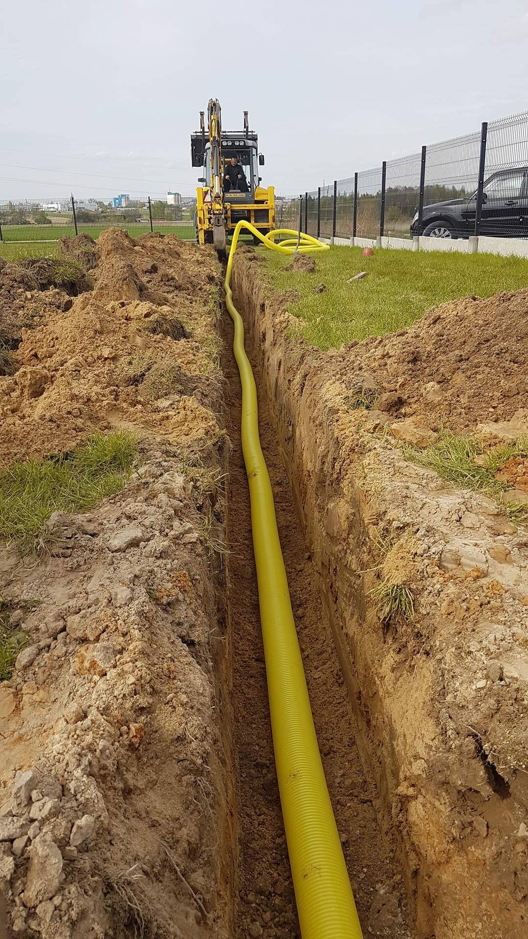 Żółta rura karbowana ułożona w wykopie ziemnym, widoczny operator koparki w tle, trawa i ogrodzenie panelowe.