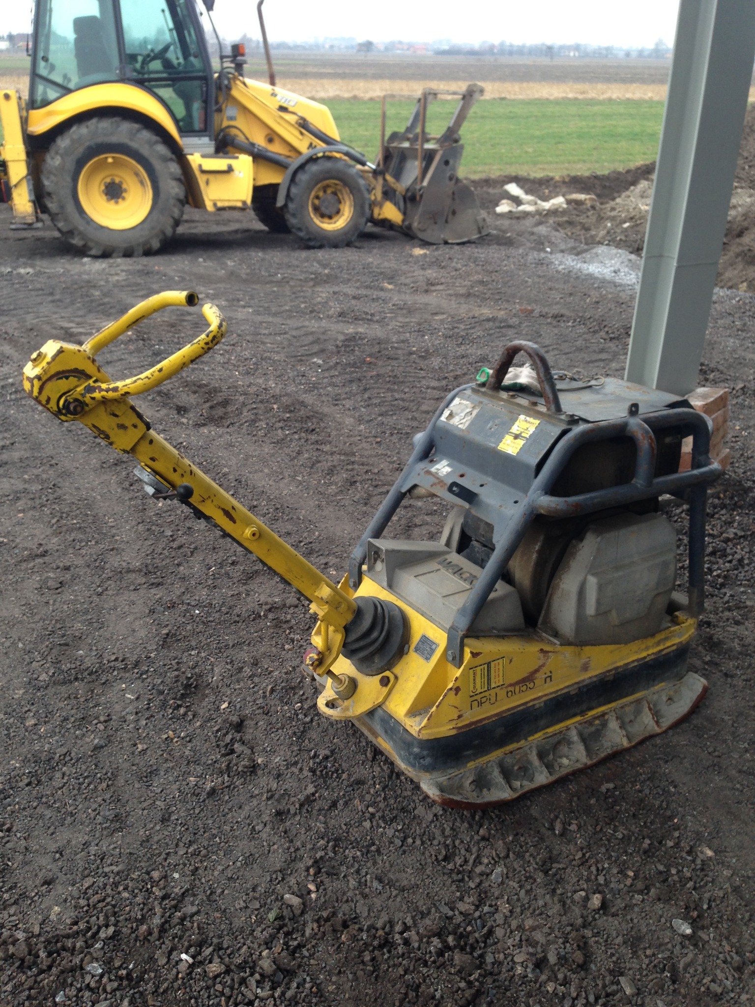 Żółta zagęszczarka wibracyjna na ubitej ziemi, z koparko-ładowarką w tle, obok metalowego słupa konstrukcyjnego.