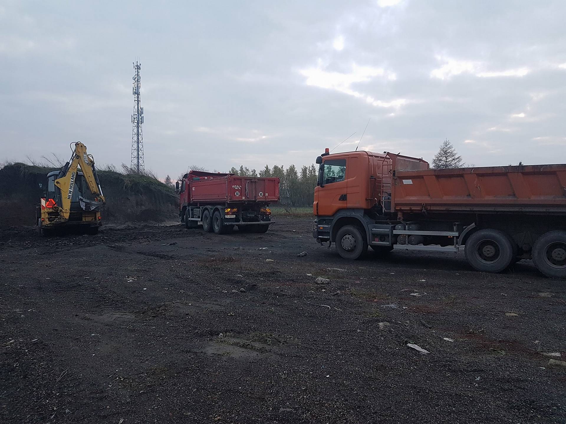 Żółta koparka New Holland i dwie wywrotki na placu budowy z ciemną ziemią, w tle maszt telekomunikacyjny i pochmurne niebo.