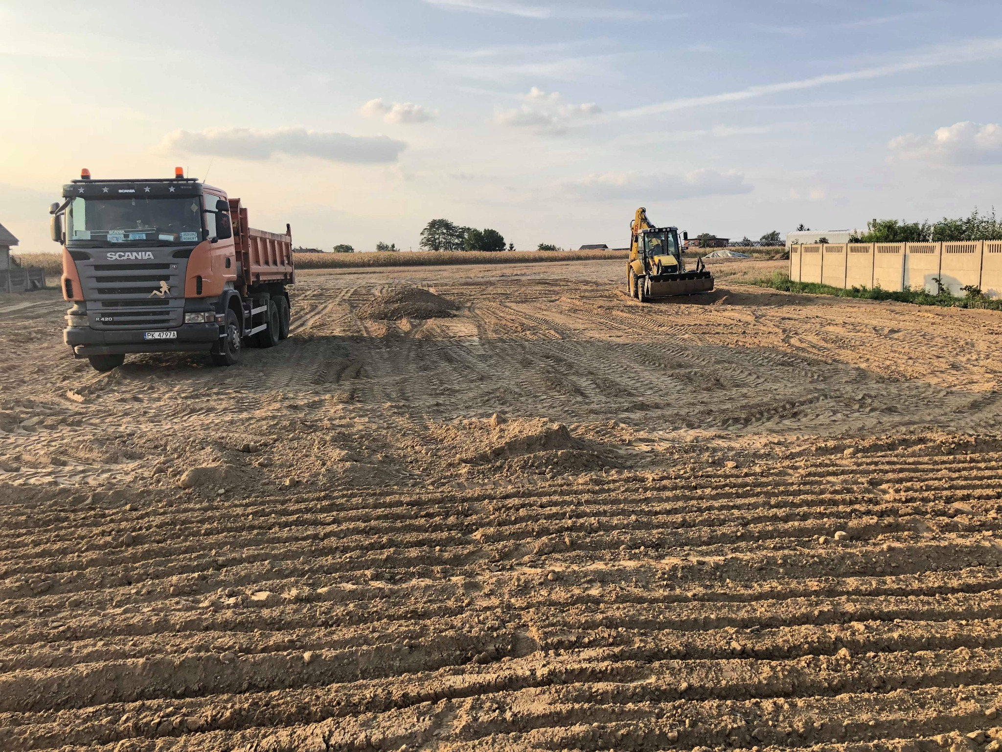 Pomarańczowa ciężarówka Scania i żółta koparka na przygotowanym terenie budowlanym z widocznymi śladami opon i świeżo usypaną ziemią.