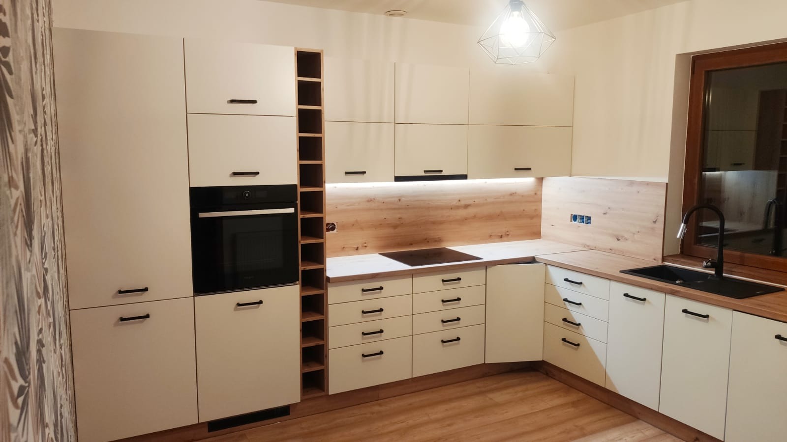 Nowoczesna kuchnia z białymi szafkami i drewnianym blatem, czarne, minimalistyczne uchwyty, zabudowany piekarnik i płyta grzewcza, drewniane panele ścienne, czarny zlew i bateria przy oknie.