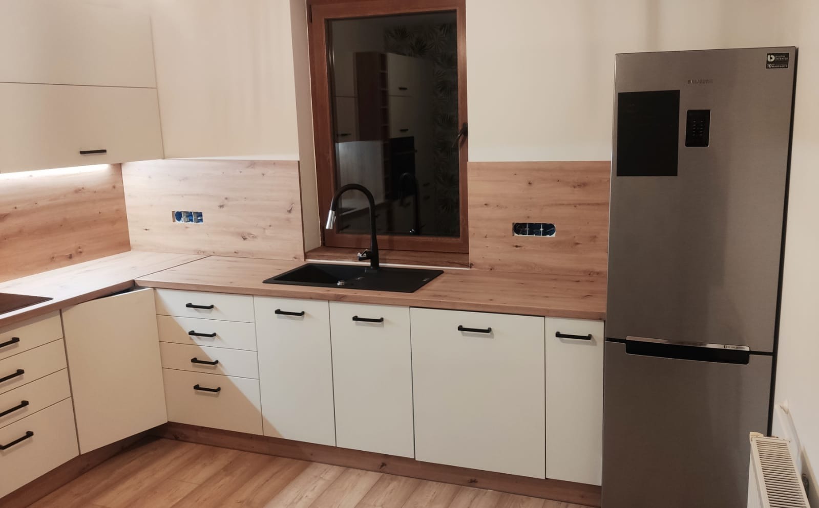 Jasna kuchnia z drewnianymi akcentami: białe szafki z czarnymi uchwytami, drewniany blat i panel nad blatem, czarny zlew i bateria, srebrna lodówka Samsung. Widoczne gniazdka elektryczne w ścianie.