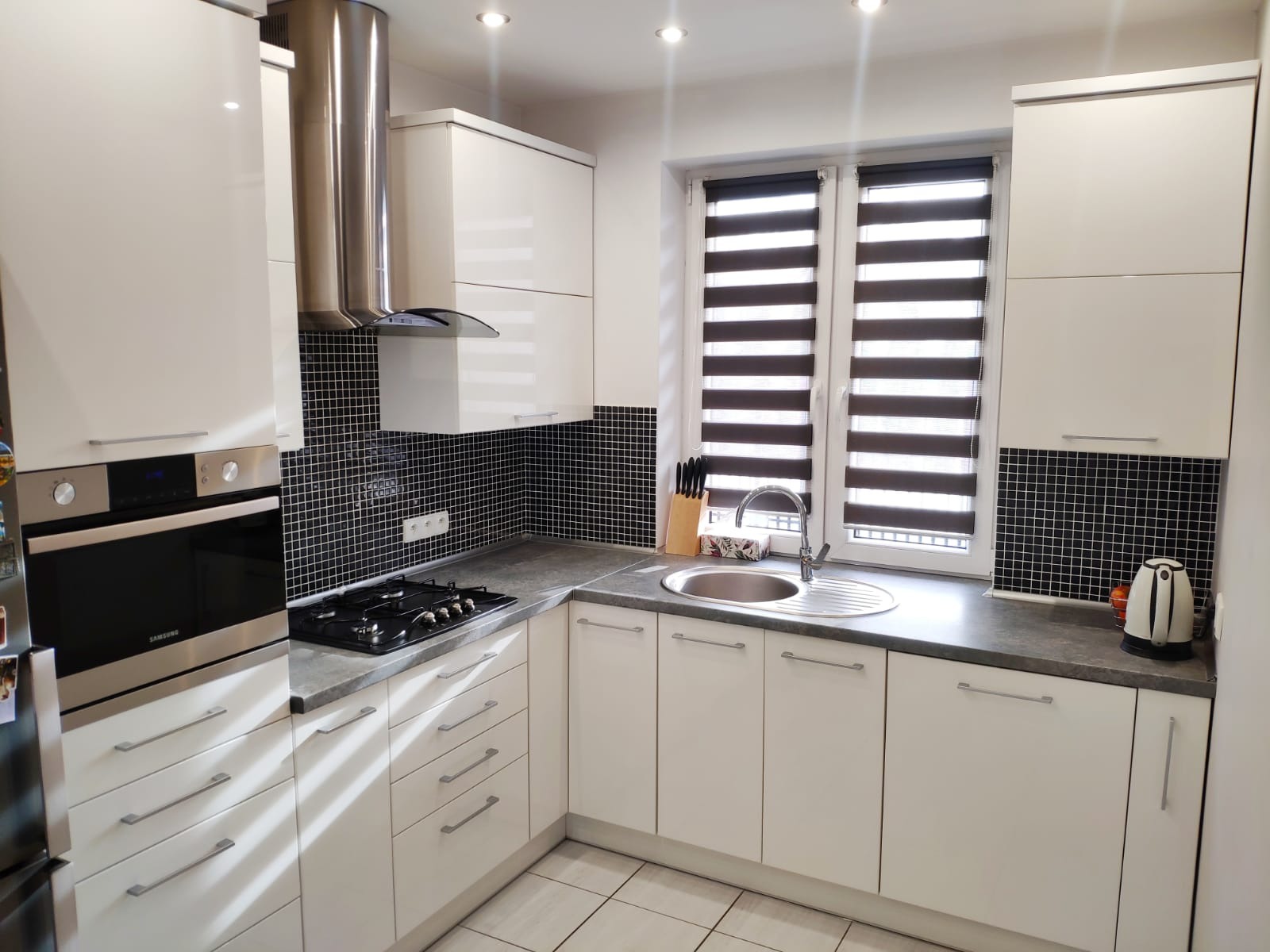 Biała kuchnia z czarną mozaiką na ścianie, stalowym okapem i roletami w oknach, widok na zlew i płytę grzewczą.