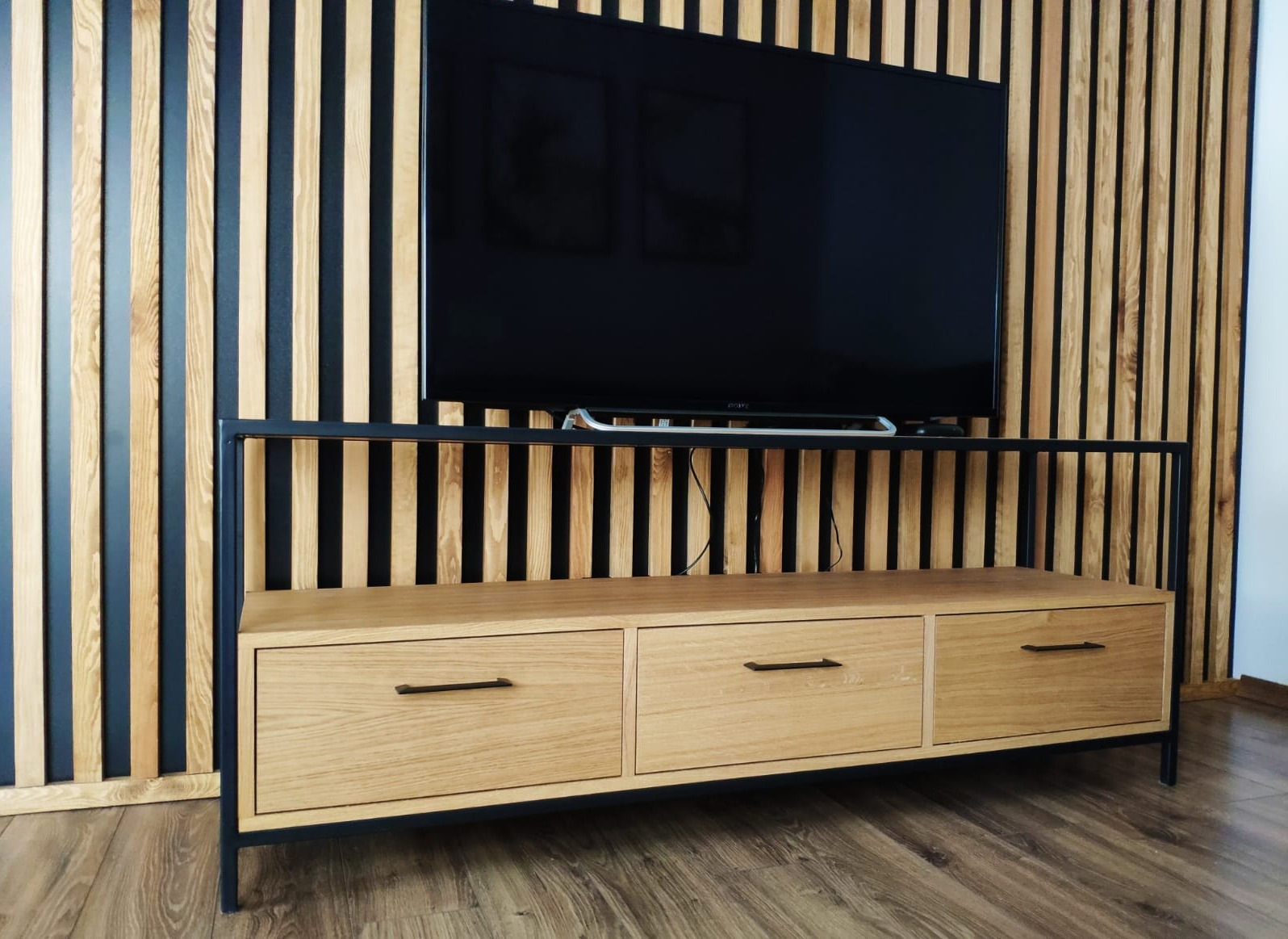 Drewniana szafka RTV z trzema szufladami i czarną metalową ramą na tle ściany z drewnianych lameli, podwieszony telewizor.