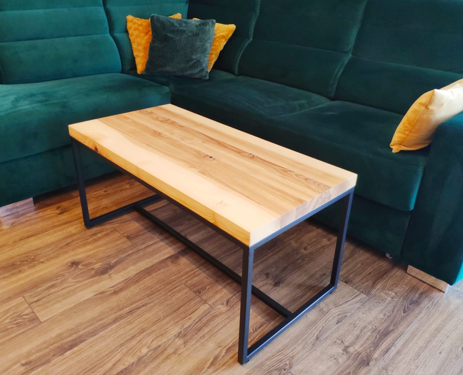 Stolik kawowy z blatem z jasnego drewna o wyraźnym usłojeniu, osadzony na czarnej, metalowej ramie, ustawiony na podłodze z paneli drewnopodobnych, obok sofy w kolorze butelkowej zieleni...