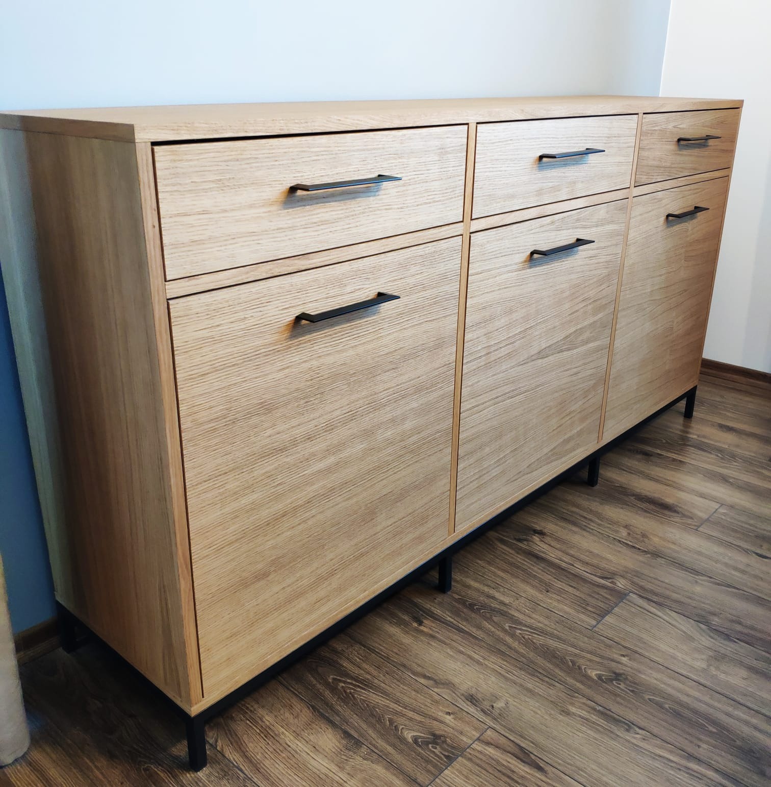 Nowoczesna komoda z jasnego drewna z czarnymi uchwytami i nóżkami, umieszczona na brązowej podłodze imitującej drewno, na tle jasnej ściany.