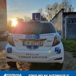 Tył białego samochodu Toyota Yaris z oznaczeniem 'L' na dachu, tablicą rejestracyjną DW 6ST58, informacją o szkoleniu na automacie i dostosowaniu pojazdu dla osób niepełnosprawnych, na tle garażu...